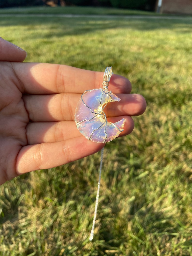 May include: A crescent moon pendant necklace. The moon-shaped pendant is made of a translucent, iridescent material, wrapped in silver-colored wire. The necklace chain is also silver-colored. The pendant is held up by a hand against a background of green grass.