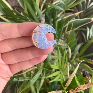 May include: A crescent moon pendant made of opalescent stone, wrapped in silver-colored wire. The pendant is held in front of a background of green and yellow-green plant leaves.