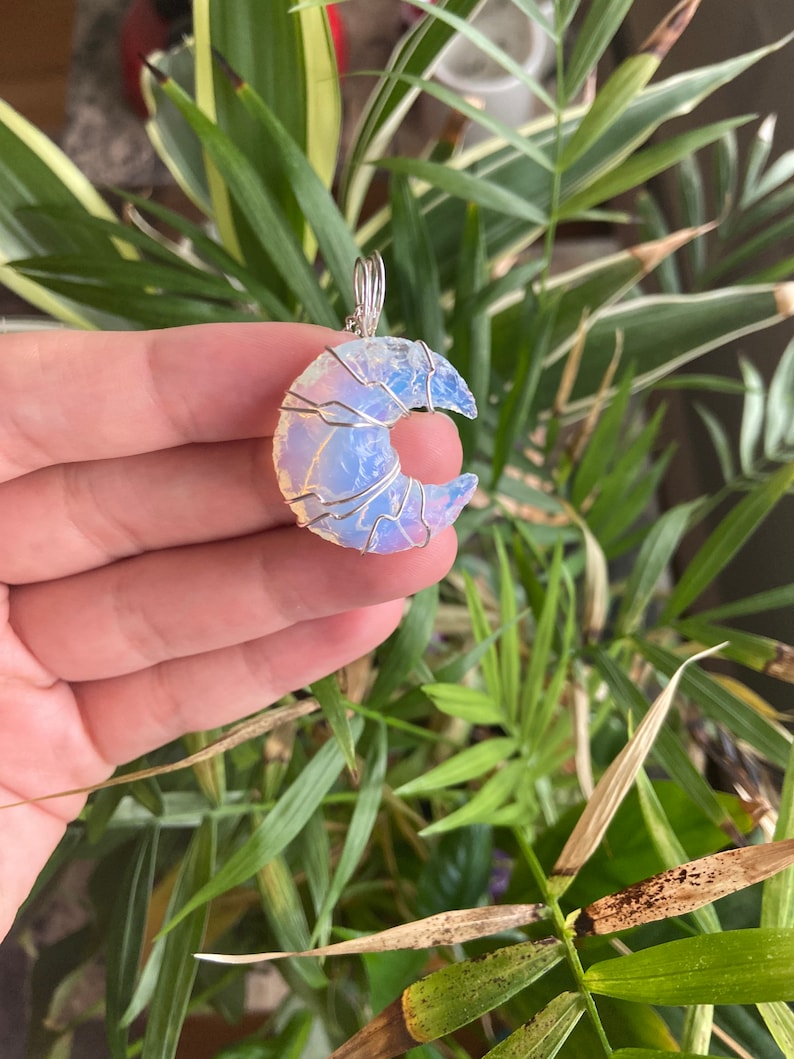 May include: A crescent moon pendant made of opalescent stone, wrapped in silver-colored wire. The pendant is held in front of a background of green and yellow-green plant leaves.