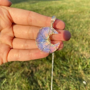 May include: A crescent moon pendant necklace. The moon-shaped pendant is made of a translucent, iridescent stone, wrapped in silver-colored wire. The necklace chain is also silver-colored. The pendant is held in a hand against a green grassy background.