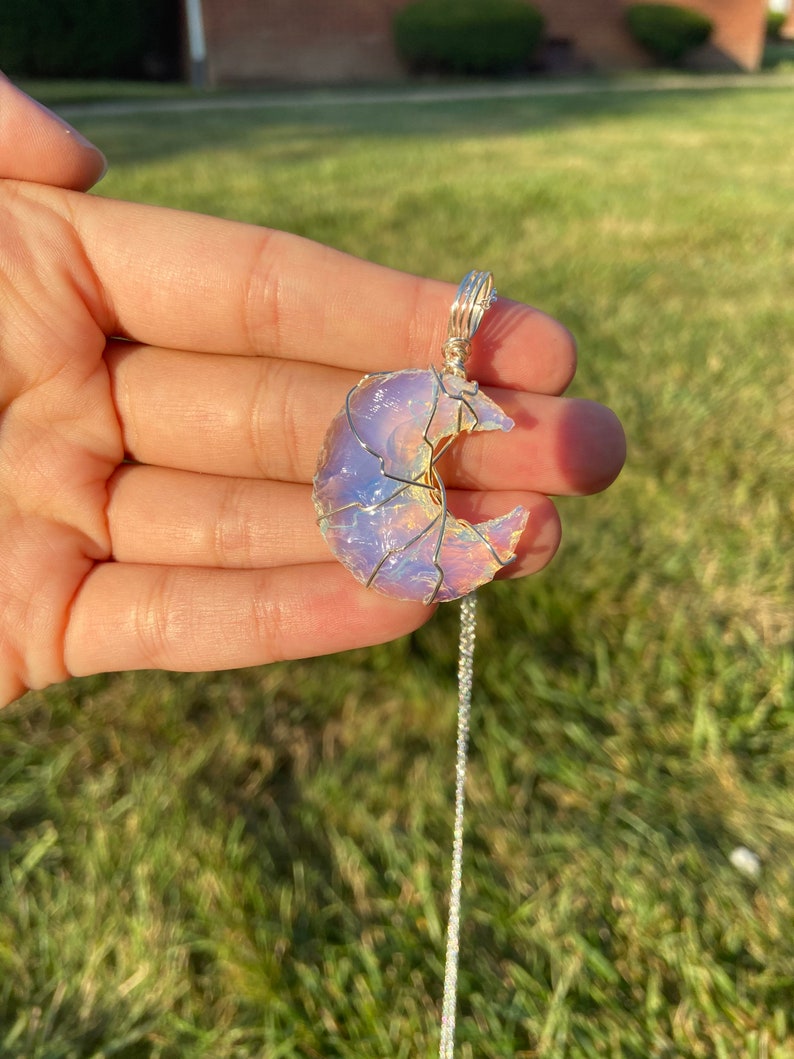 May include: A crescent moon pendant necklace. The moon-shaped pendant is made of a translucent, iridescent stone, wrapped in silver-colored wire. The necklace chain is also silver-colored. The pendant is held in a hand against a green grassy background.