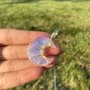 May include: A crescent moon pendant with a silver wire wrap and chain. The moon is a translucent, iridescent stone with a soft, pearly glow. The pendant is held in a hand against a blurred green background.