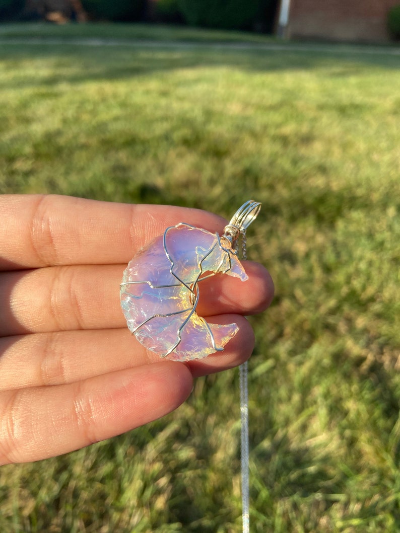 May include: A crescent moon pendant with a silver wire wrap and chain. The moon is a translucent, iridescent stone with a soft, pearly glow. The pendant is held in a hand against a blurred green background.