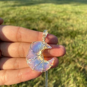 May include: A crescent moon pendant with a silver wire frame and a shimmering, opalescent stone. The pendant hangs from a delicate silver chain, creating a celestial-themed necklace.