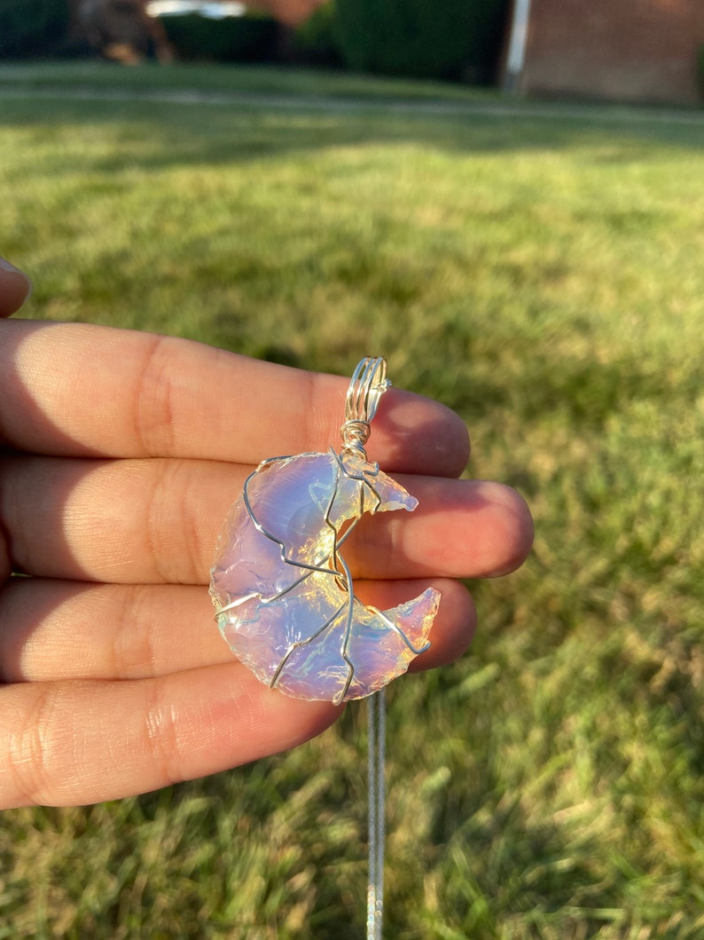 May include: A crescent moon pendant with a silver wire frame and a shimmering, opalescent stone. The pendant hangs from a delicate silver chain, creating a celestial-themed necklace.
