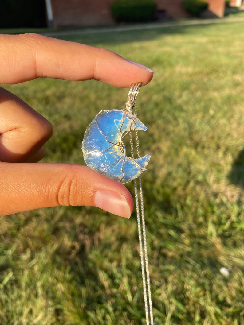May include: A silver necklace featuring a crescent moon pendant. The moon is crafted from a light blue, translucent stone, wrapped in silver wire. The necklace hangs on a delicate silver chain, creating a celestial-themed accessory.