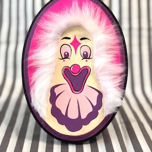 May include: Oval-shaped clown face with a cream-colored face, pink cheeks, and a purple and pink ruffled collar. The clown has a pink diamond on its forehead, large eyes, and a wide, open mouth. The face is framed by a fluffy white wig and a pink and purple border.