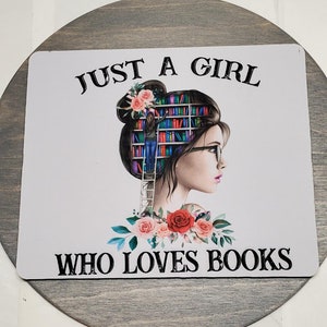 May include: A white sign with the text "Just a girl who loves books" in black lettering. The sign features a colorful illustration of a woman with a bookshelf in her head and flowers around her neck.