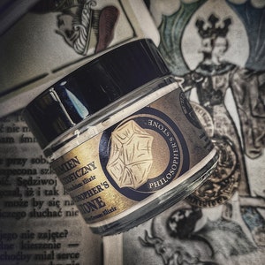 May include: A glass jar with a black lid, labeled "Philosopher's Stone" and "Elixir". The label features a gold emblem and text. The jar is positioned near playing cards and a vintage illustration.