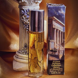 May include: A clear glass roll-on perfume bottle with a silver cap, filled with a golden liquid and glitter. The bottle is next to a box with an illustration of a building with columns. The text on the box reads "Ancient Perfume Collection Library of Alexandria Roll-on 10 ml 0.33 FL OZ."