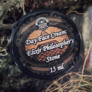 Könnte beinhalten: Ein schwarzer Tiegel mit einem gold-schwarzen Etikett, auf dem "Day Face Cream: Elixir Philosopher's Stone 15 ml" steht. Der Tiegel liegt auf einem Bett aus braunen Wurzeln und Moos.