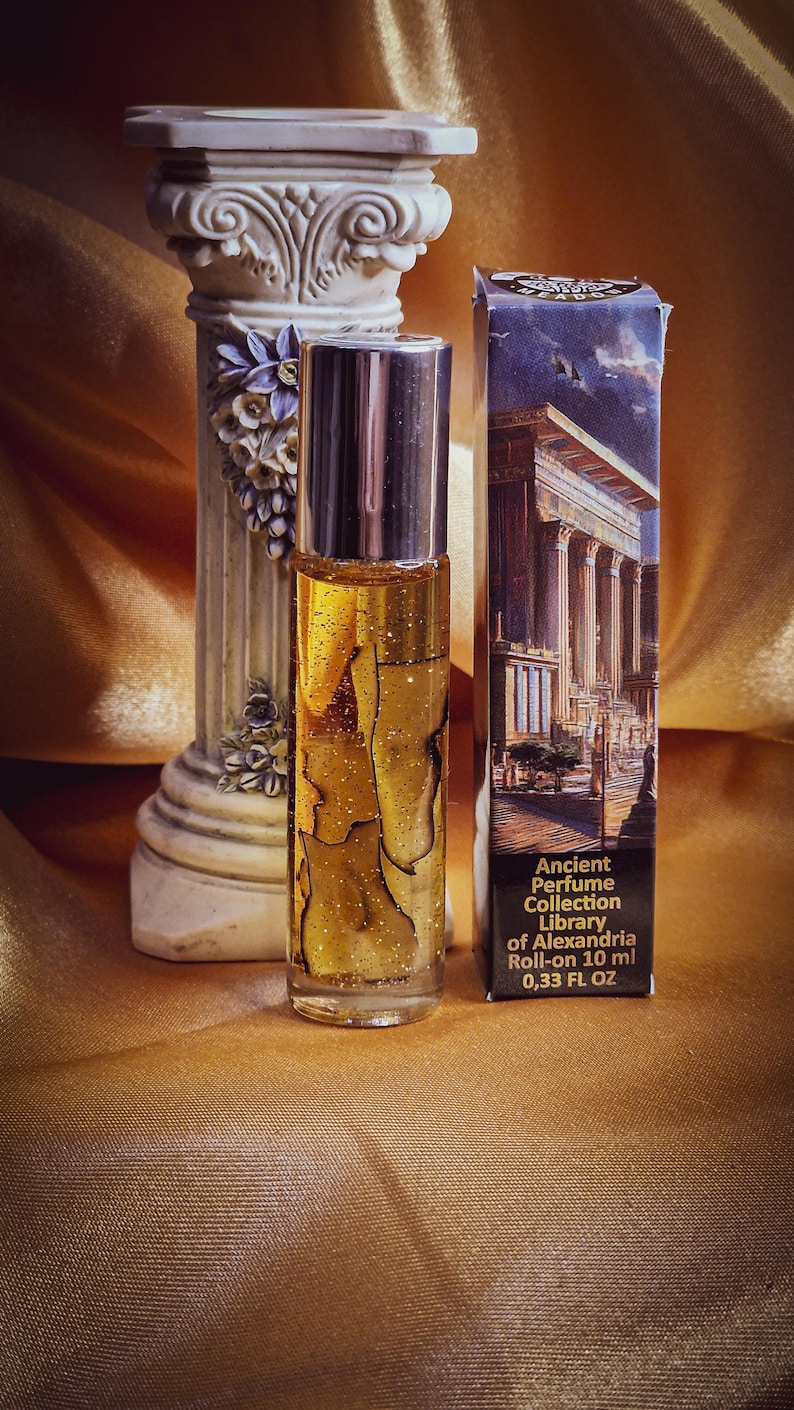 May include: A clear glass roll-on perfume bottle with a silver cap, filled with a golden liquid and glitter. The bottle is next to a box with an illustration of a building with columns. The text on the box reads "Ancient Perfume Collection Library of Alexandria Roll-on 10 ml 0.33 FL OZ."