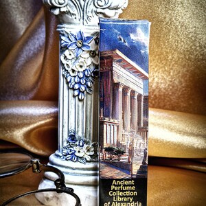 May include: A decorative white column with blue and white floral accents stands next to a perfume box. The box features an illustration of an ancient building and reads "Ancient Perfume Collection Library of Alexandria Roll-on 10 ml 0.33 FL OZ."