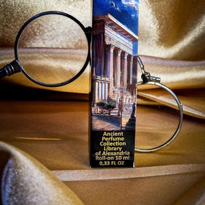 May include: A perfume roll-on bottle with a label featuring an illustration of an ancient building. The label reads "Ancient Perfume Collection Library of Alexandria Roll-on 10 ml 0.33 FL OZ". The bottle is set against a gold fabric background.