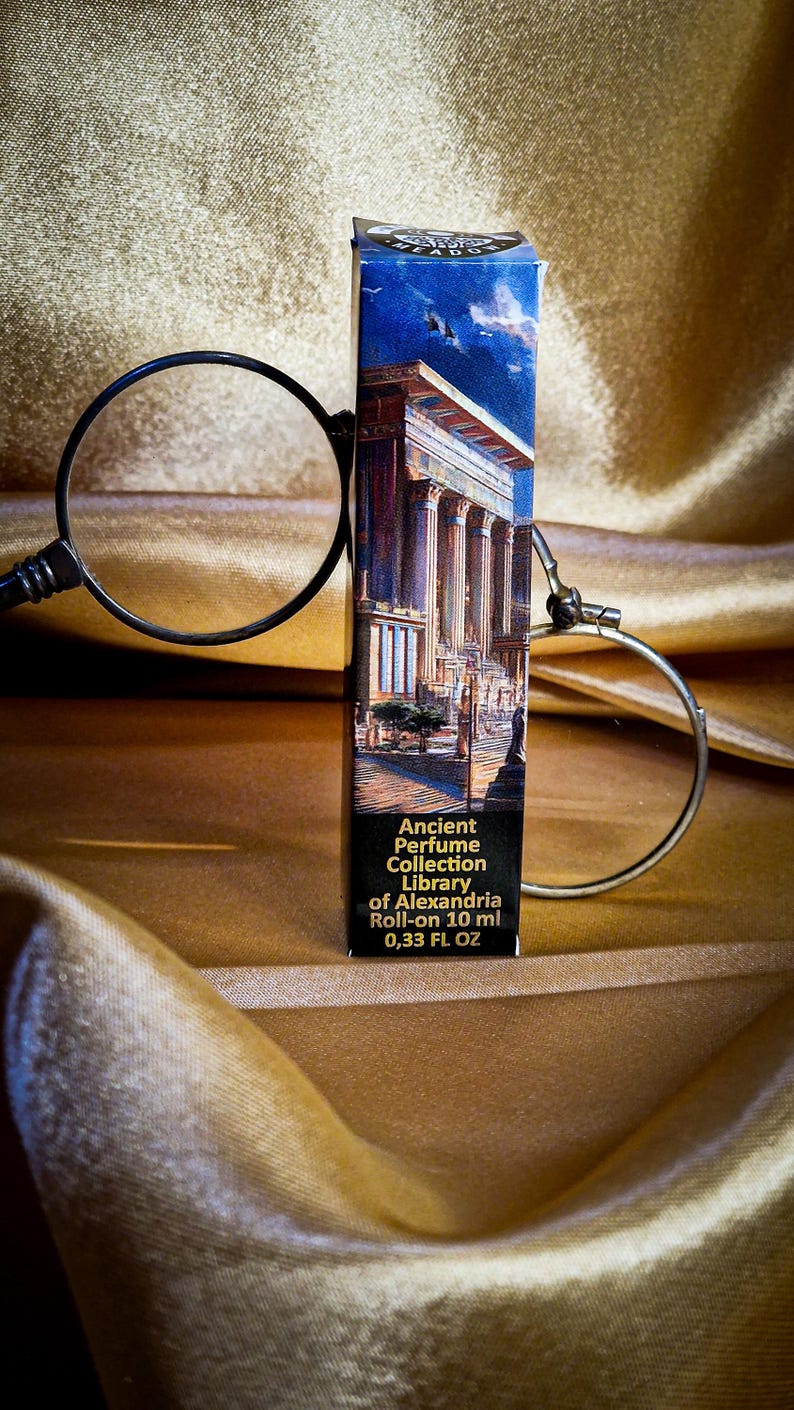 May include: A perfume roll-on bottle with a label featuring an illustration of an ancient building. The label reads "Ancient Perfume Collection Library of Alexandria Roll-on 10 ml 0.33 FL OZ". The bottle is set against a gold fabric background.