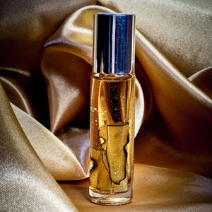 May include: A clear glass perfume bottle with a silver cap, filled with a golden liquid. The bottle is set against a backdrop of shimmering, golden fabric. The liquid contains a dark, abstract design.