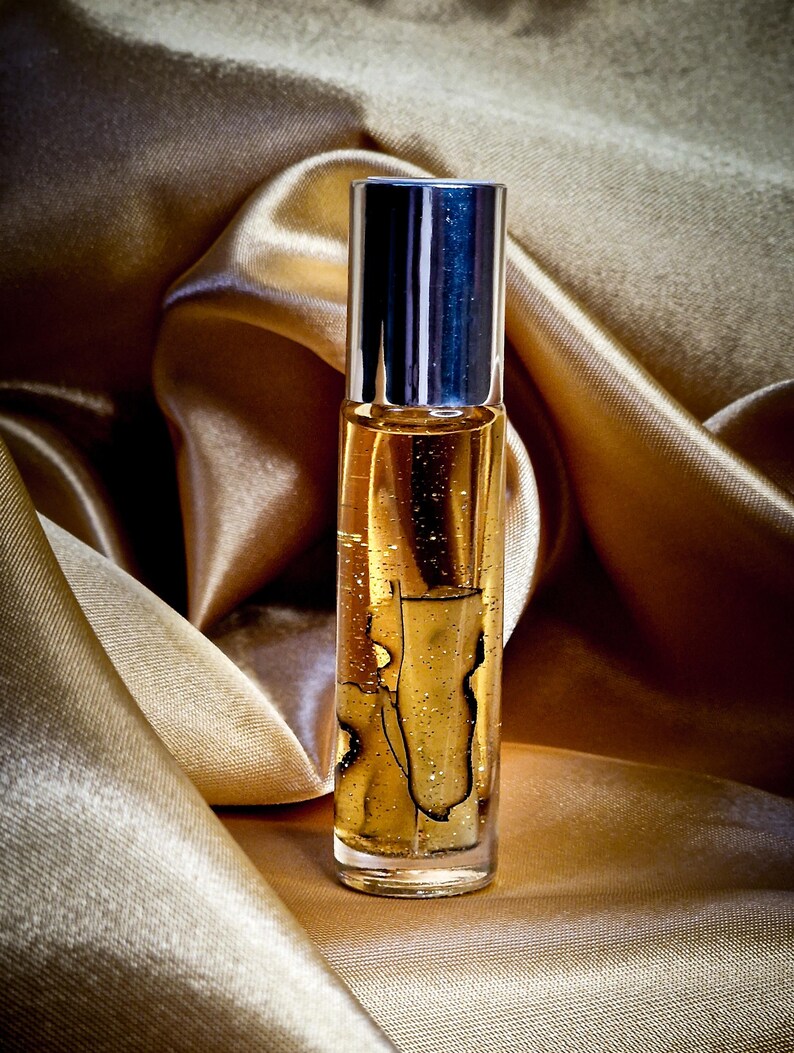 May include: A clear glass perfume bottle with a silver cap, filled with a golden liquid. The bottle is set against a backdrop of shimmering, golden fabric. The liquid contains a dark, abstract design.