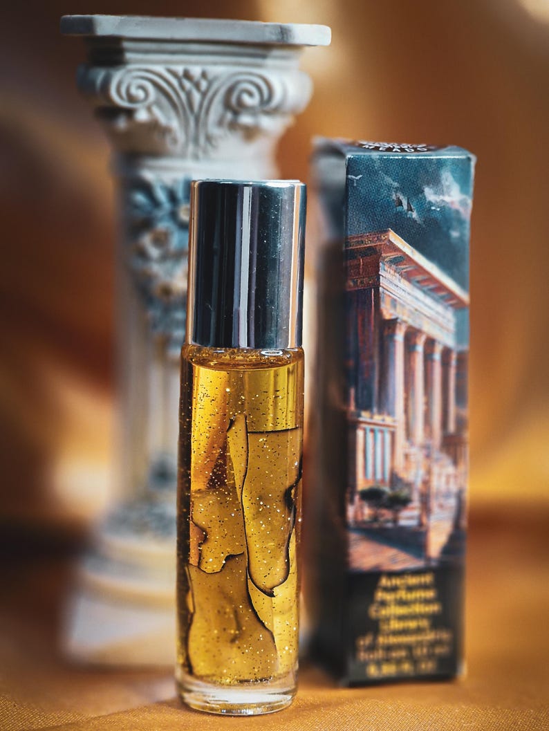 May include: A clear glass perfume bottle with a silver cap, filled with a golden liquid and glitter. The bottle is next to a box with a classical building illustration and a decorative column. The background is a warm, golden color.