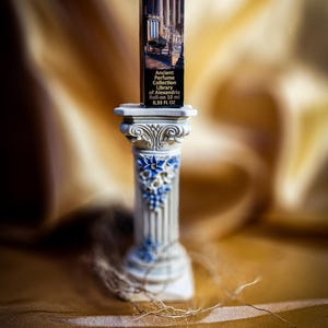 May include: A small, rectangular perfume bottle with a classical building illustration, labeled "Ancient Perfume Collection Library of Alexandria". The bottle is displayed on a decorative, white and blue column. The background is a blurred, golden fabric.