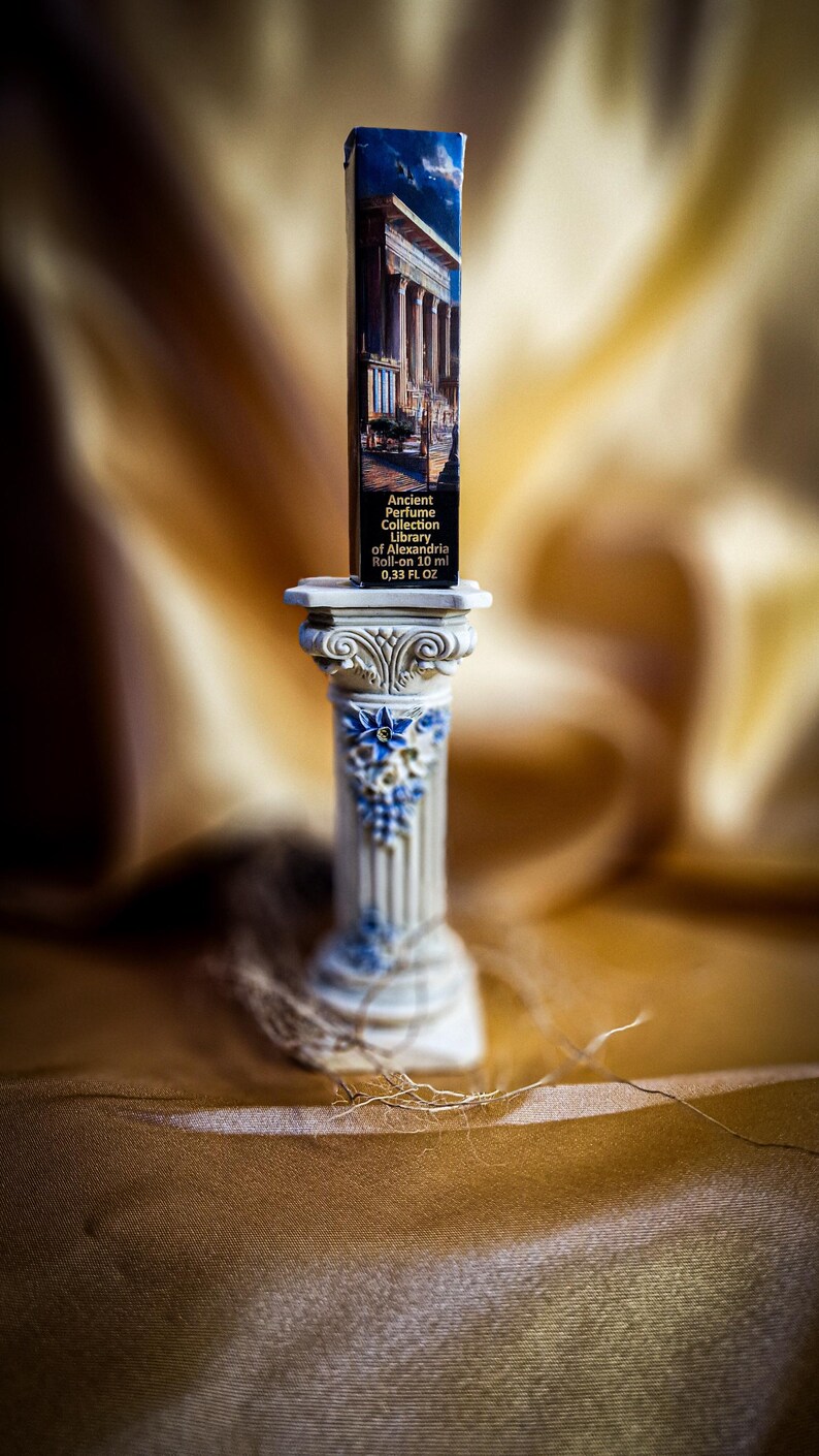 May include: A small, rectangular perfume bottle with a classical building illustration, labeled "Ancient Perfume Collection Library of Alexandria". The bottle is displayed on a decorative, white and blue column. The background is a blurred, golden fabric.