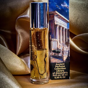 May include: A roll-on perfume bottle with a silver cap and a gold-colored liquid, next to a box with a classical building illustration. The box reads "Ancient Perfume Collection Library of Alexandria Roll-on 10 ml 0.33 FL OZ". The background is a gold satin fabric.