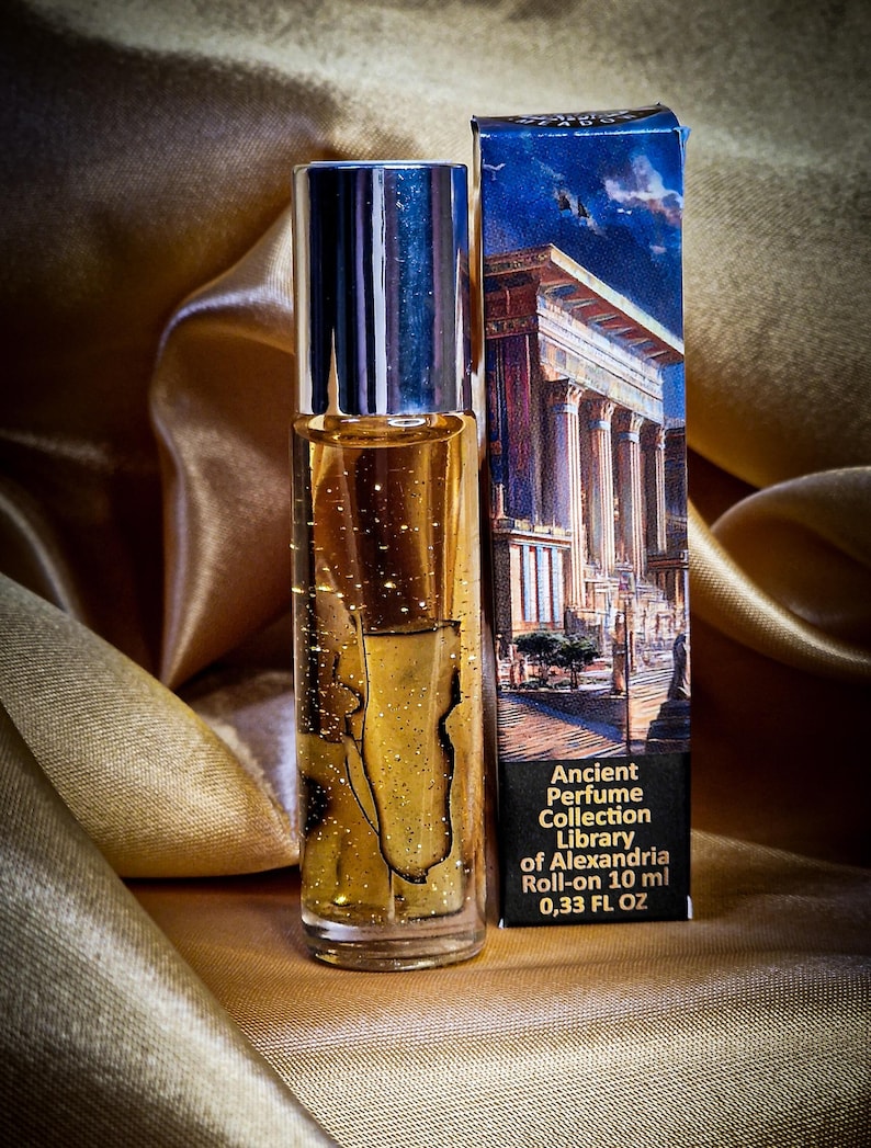 May include: A roll-on perfume bottle with a silver cap and a gold-colored liquid, next to a box with a classical building illustration. The box reads "Ancient Perfume Collection Library of Alexandria Roll-on 10 ml 0.33 FL OZ". The background is a gold satin fabric.