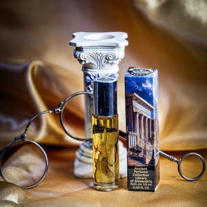 May include: A perfume bottle with a black cap and gold liquid, next to a blue and white box with the text "Ancient Perfume Collection Library of Alexandria". A decorative column and a pair of antique spectacles are also in the image.