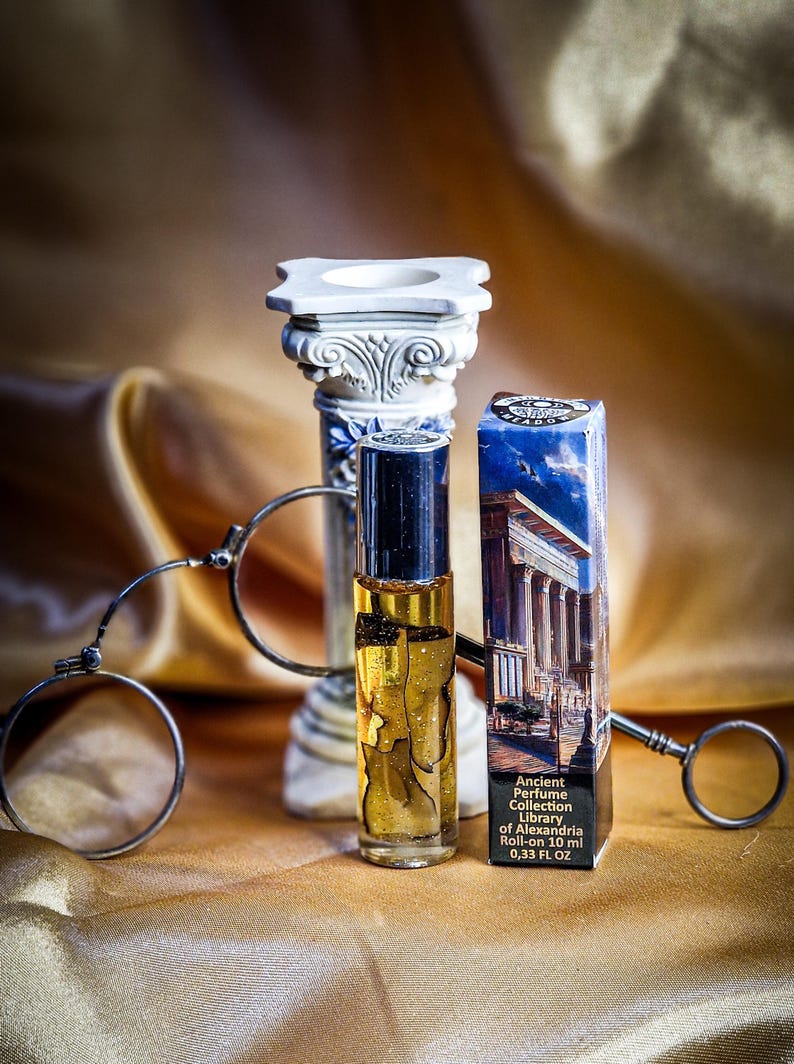 May include: A perfume bottle with a black cap and gold liquid, next to a blue and white box with the text "Ancient Perfume Collection Library of Alexandria". A decorative column and a pair of antique spectacles are also in the image.