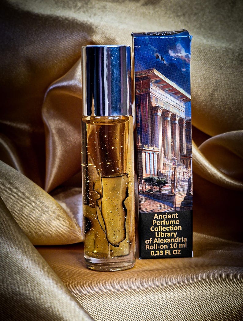 May include: A roll-on perfume bottle with a silver cap and a gold-colored liquid, next to a box with a classical building illustration. The box reads "Ancient Perfume Collection Library of Alexandria Roll-on 10 ml 0.33 FL OZ". The background is a gold satin fabric.