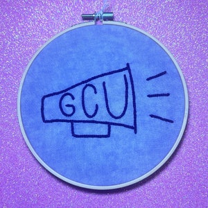 May include: A blue embroidery hoop with a white background and a black embroidered megaphone with the text "GCU" inside.