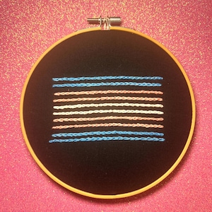 Puede incluir: Un aro de bordado negro con un diseño de bandera del orgullo transgénero bordado en hilo azul, rosa y blanco.