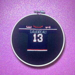 May include: A black and white cross-stitch embroidery of a football jersey with the name "GAULDREAU" and the number "13" on it. The embroidery is framed in a black and white hoop.