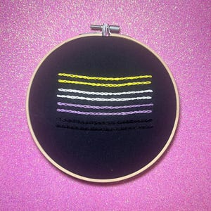 May include: A black embroidery hoop with a multi-colored embroidery design. The design features five horizontal lines in yellow, white, purple, and black.