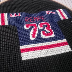 May include: Cross-stitch embroidery of a blue hockey jersey with the name "REMPE" and the number "73" in red and white. The jersey is set against a black background within a wooden embroidery hoop. The design features red and white stripes.