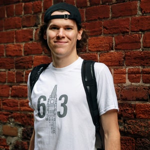 May include: A person wearing a white t-shirt with a black and white graphic of a building and the number 613. The person is wearing a black baseball cap and a black backpack.