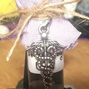 May include: A small glass bottle with a silver medical symbol charm, wrapped in a lavender fabric and tied with twine.