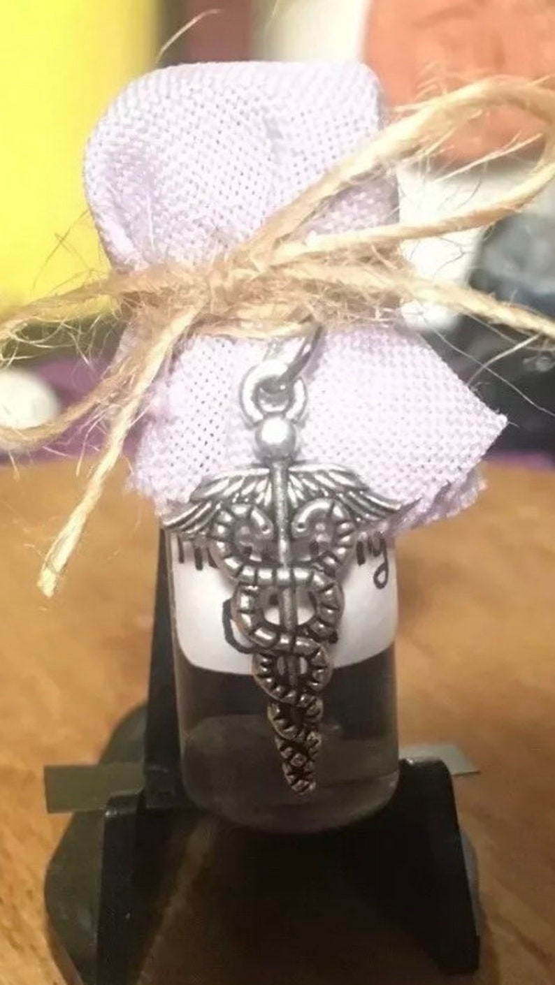 May include: A small glass bottle with a silver medical symbol charm, wrapped in a lavender fabric and tied with twine.