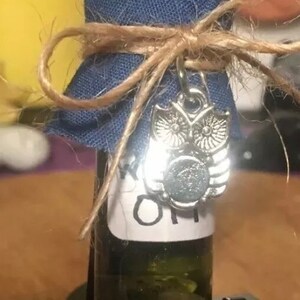 May include: A small glass bottle with a blue fabric cover tied with twine and a silver owl charm. The bottle is filled with a clear liquid and has a white label that says "Sage Oil".