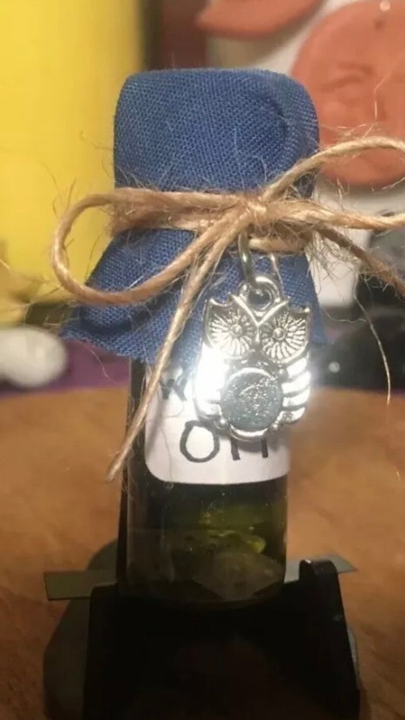 May include: A small glass bottle with a blue fabric cover tied with twine and a silver owl charm. The bottle is filled with a clear liquid and has a white label that says "Sage Oil".