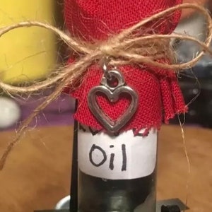 May include: A small glass bottle with a red fabric cover tied with twine and a silver heart charm. The bottle is labeled "oil".