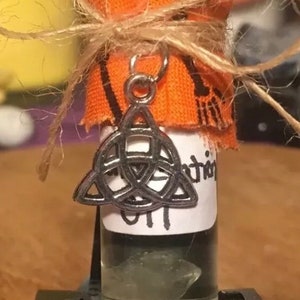 May include: A small glass bottle with a clear liquid inside. The bottle is wrapped in orange fabric and tied with twine. A silver charm in the shape of a triquetra hangs from the bottle.