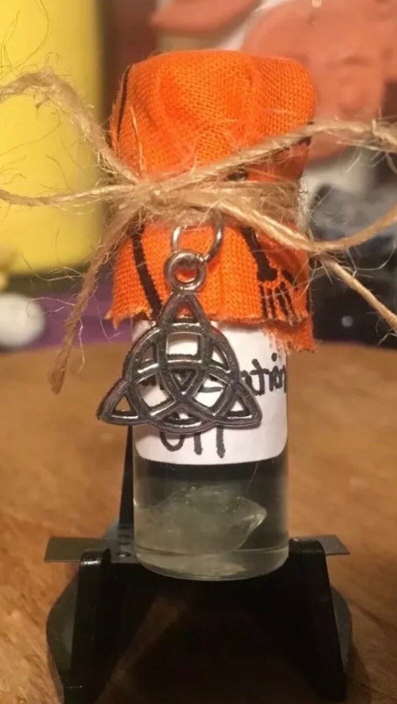 May include: A small glass bottle with a clear liquid inside. The bottle is wrapped in orange fabric and tied with twine. A silver charm in the shape of a triquetra hangs from the bottle.