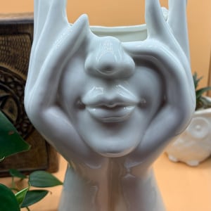 May include: A white ceramic planter shaped like a face with hands framing the face. The planter has a smiling mouth and a nose. The hands are raised, with the fingers pointing upwards. The planter is a decorative item.