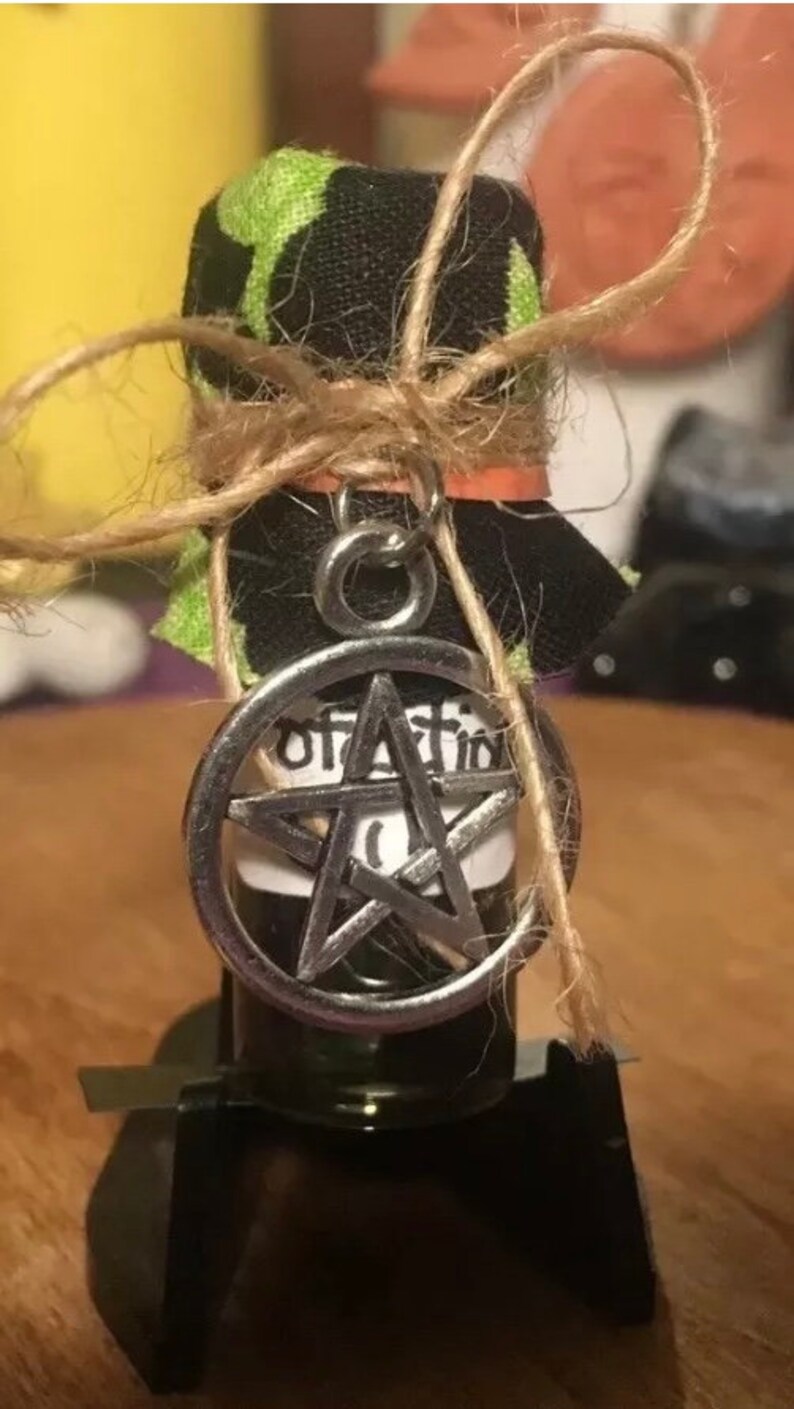 May include: A small glass bottle with a black and green fabric cover tied with twine. The bottle has a silver pendant with a pentagram charm and the word "protection" written on the label.