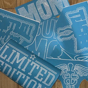 May include: A collection of blue stencils with white dot patterns, laid out on a wooden surface. The stencils display the words "MOM", "LIMITED EDITION", and other designs. These stencils are likely for crafting or DIY projects.