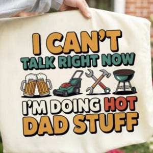 May include: A cream-colored tote bag featuring the text I CAN'T TALK RIGHT NOW I'M DOING HOT DAD STUFF in yellow, green, and red. The design includes illustrations of two beer mugs, a green lawnmower, crossed metal tools, and a black charcoal grill.