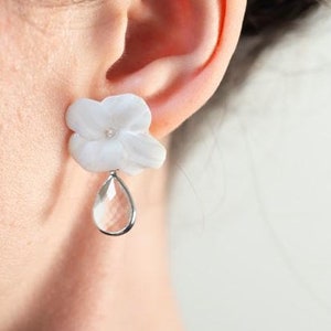 May include: White flower-shaped earrings with a clear teardrop-shaped charm. The earrings are made of a white material and feature a small, clear gem in the center of the flower. The teardrop charm hangs from a silver-colored ring. The earrings are worn on the ear.