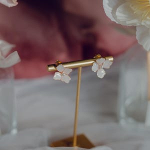 Könnte beinhalten: Ein Paar zarte Blumenohrringe auf einem goldenen Präsentationsständer. Die Ohrringe haben weiße Blütenblätter mit einem Perlenmittelpunkt. Der Hintergrund umfasst weiche rosa und weiße florale Elemente.