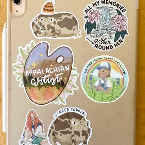 May include: A collection of stickers featuring various illustrations, including a frog wearing a hat, a frog with a grumpy expression, a watercolor palette with the words "Appalachian artist", a scene with a waterfall and flowers, and a person gardening. The stickers are on a light brown surface.