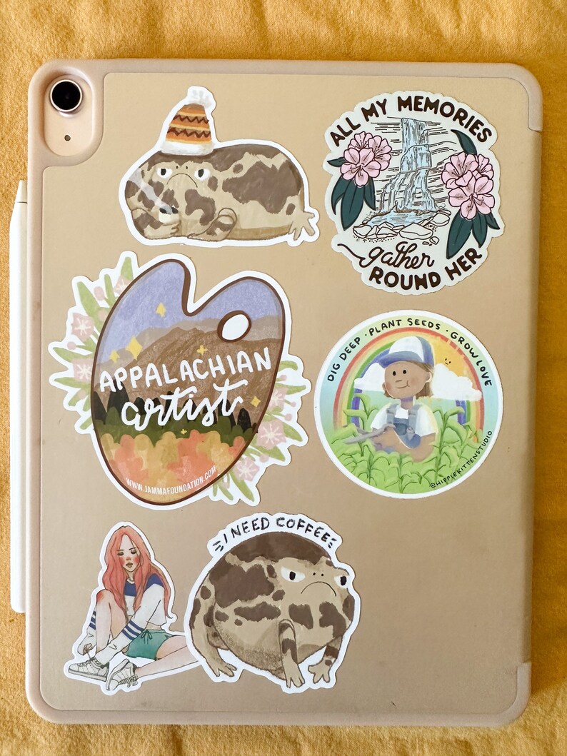 May include: A collection of stickers featuring various illustrations, including a frog wearing a hat, a frog with a grumpy expression, a watercolor palette with the words "Appalachian artist", a scene with a waterfall and flowers, and a person gardening. The stickers are on a light brown surface.
